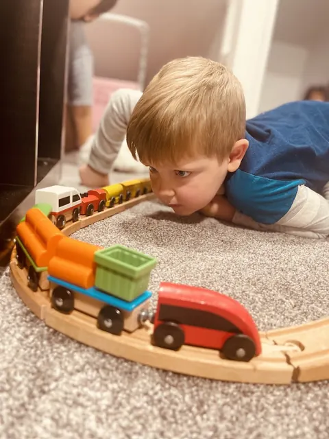 A toddler lying on the carpet, eye level with a wooden toy train on its track