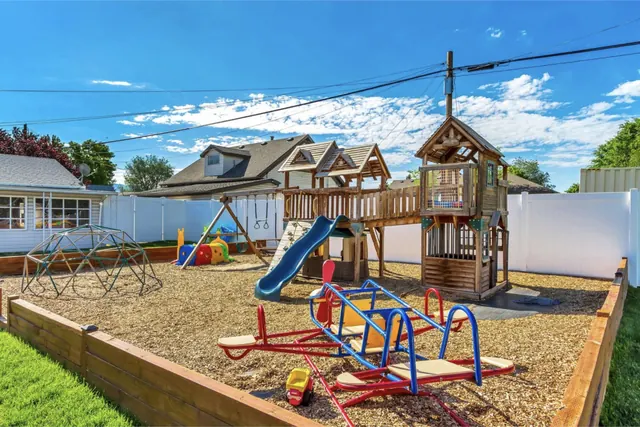 Cari’s backyard playground: a wooden playset with a slide and swings, a dome climber, a seesaw and a timber-edged sandbox under a blue sky